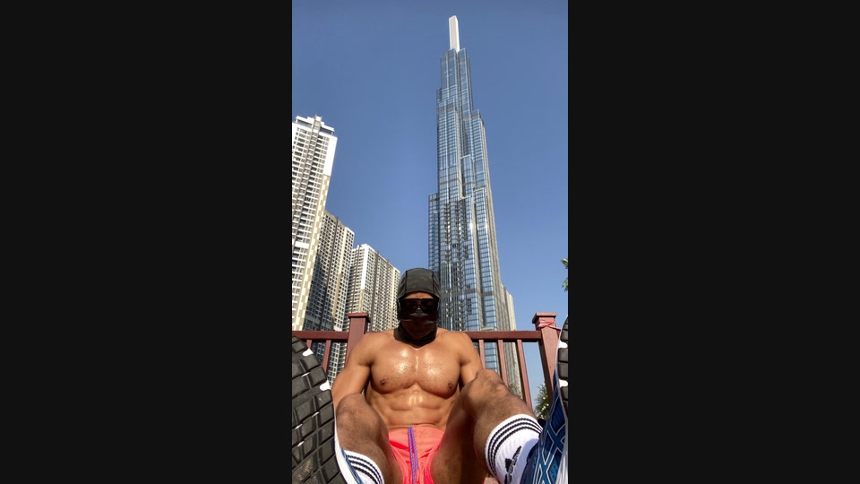 Low-angle daylight photograph of a masked athlete seated outdoors, with high-rise buildings behind him.