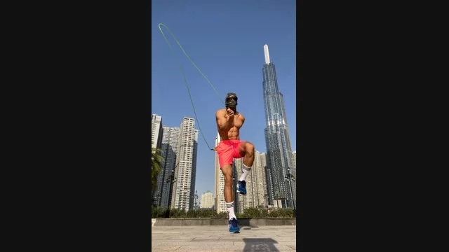 Masked athlete jumping rope outdoors in daylight, framed by high-rise buildings and a clear blue sky.