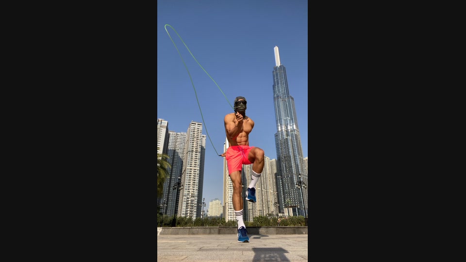 Masked athlete jumping rope outdoors in daylight, framed by high-rise buildings and a clear blue sky.