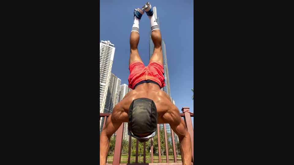 Close low-angle view of a masked athlete inverted during an outdoor handstand, with buildings and blue sky behind him.